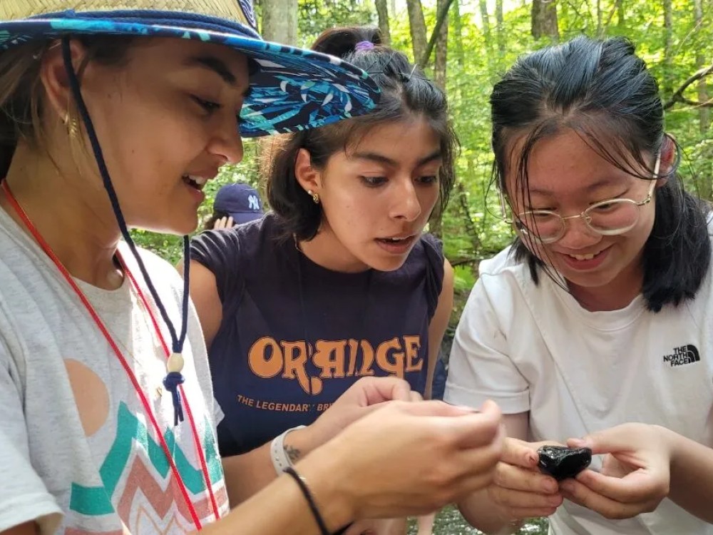 Program leader and two students looking at a rock out in the field in a wooded area